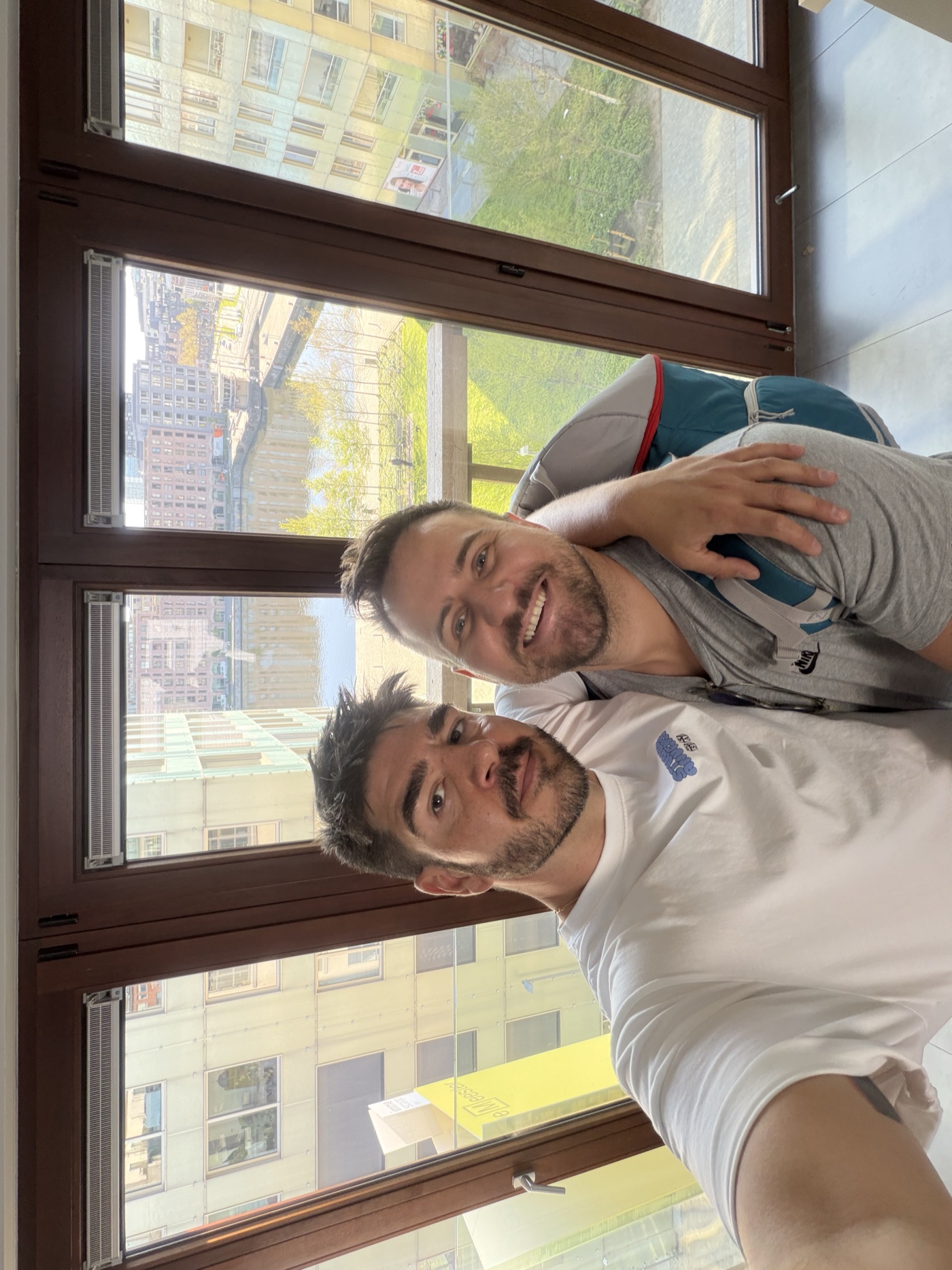 Daan Vervoort y Daniel Moncau, sonrientes y abrazados, se hacen un selfi junto a un gran ventanal con vistas a un canal y edificios.