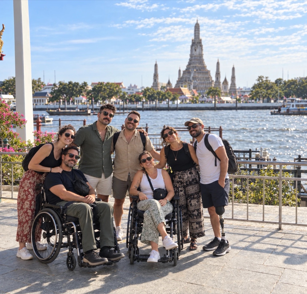 Un grupo de seis amigos, dos de ellos usuarios de silla de ruedas y uno con prótesis de pierna, posan sonrientes junto a un río con un templo al fondo.